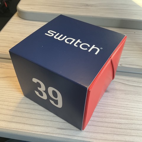 Würfelbox, zwei dunkelblaue Seiten sind zu sehen, auf einer steht weiss „Swatch“, auf der anderen „39“, eine rote Seite