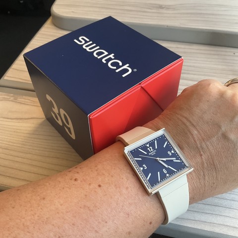 gleiche Box, davor ein linkes Handgelenk mit der Uhr: beiges Band und Gehäuse, Zifferblatt quadratisch dunkelblau mit weisser Beschriftung und Zeigern