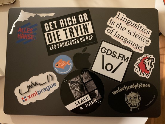 stickers on laptop case: Fischland-Darß-Zingst, Hanse, “Get rich or die trying'”, “Lingusitics is the science of langauge”, GDS.FM, LHC, Motörheadphönes, ZHAW Digital, XMLPrague