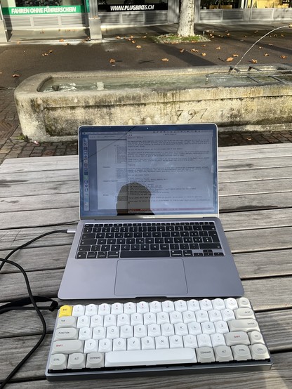 Photo of a MacBook and a mechanical keyboard on an outdoor table in front of a fountain with the inscription “1797.”
