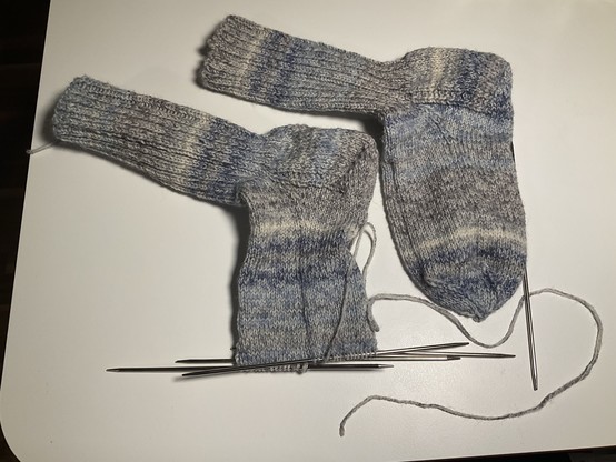 zwei graumelierte Socken, der linke ist noch nicht fertig, Nadeln sind noch drin, Fussspitze fehlt, Ende des Wollfadens ist aber schon da