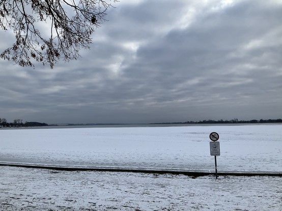 grauer See mit Schnee bedeckt unter grauem Himmel. Vorne ein Schild zweigeteilt: oben Schwimmen verboten, unten Eisfläche gesperrt Einbruchgefahr

Unteruckersee