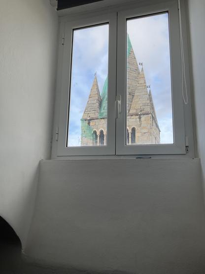 weisse Wand, oben ein geschlossenes doppelflügeliges Fenster, durch das man Kirchturm und blauen Himmel sieht 